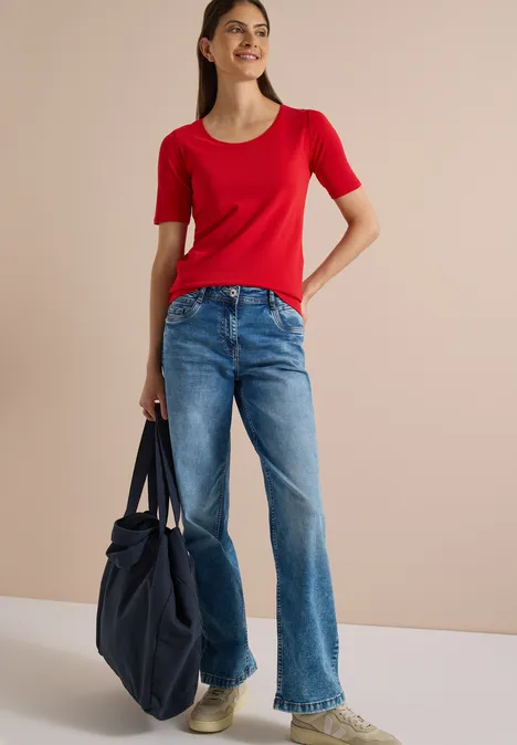 Frau trägt rotes Oberteil, blaue Jeans und hält eine große blaue Tasche.