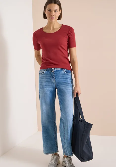 Frau trägt rotes T-Shirt, blaue Jeans und hält eine dunkelblaue Tasche.
