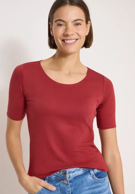 Junge Frau trägt rotes kurzärmeliges T-Shirt und blaue Jeans vor hellem Hintergrund.