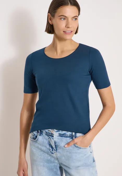 Frau trägt dunkelblaues kurzärmeliges Shirt und helle Jeans, eine Hand in Tasche, neutraler Hintergrund
