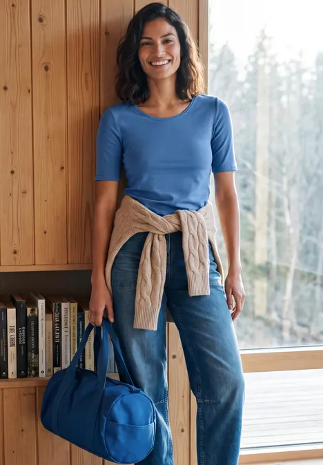 Lächelnde Frau in blauem Shirt und Jeans, hält eine blaue Tasche vor Fenster und Bücherregal.