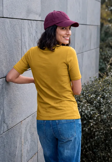 Basic T-shirt met ronde hals golden honey Frau im gelben T-Shirt, Jeans und lila Cap lehnt an einer Mauer und blickt lächelnd über die Schulter.