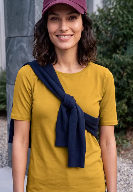 Basic T-shirt met ronde hals golden honey Eine lächelnde Frau in gelbem T-Shirt, mit dunkelblauem Pullover um die Schultern und rosa Cap.