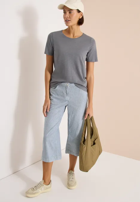 Basic Shirt met ronde hals graphite light grey Vrouw in grijs T-shirt, lichtblauwe cropped jeans, witte sneakers en pet, met kaki tas tegen lichte achtergrond.