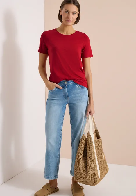 Basic T-Shirt mit Rundhals in Unifarbe tamarillo red Frau trägt rotes T-Shirt, blaue Jeans und hält Strohtasche vor zweifarbigem Hintergrund.