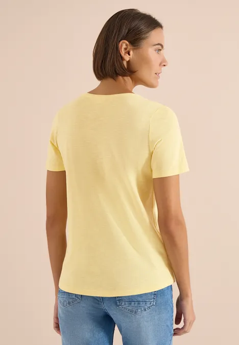 Eine Person steht mit dem Rücken zur Kamera und trägt ein gelbes T-Shirt und blaue Jeans.