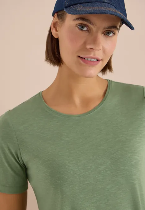 Person mit grünem T-Shirt und blauem Cap vor neutralem Hintergrund.