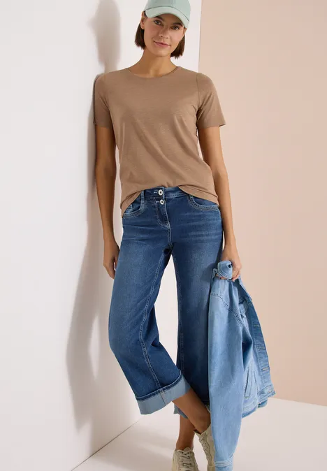 Basic T-Shirt mit Rundhals in Unifarbe creamy mocha Frau lehnt an Wand, trägt braunes T-Shirt, blaue Jeans und hält Jeansjacke in der Hand.