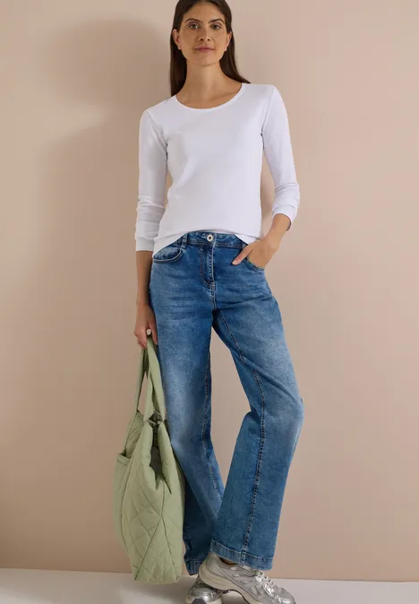 Frau trägt weißes Langarmshirt, blaue Jeans und hält eine gesteppte grüne Tasche.