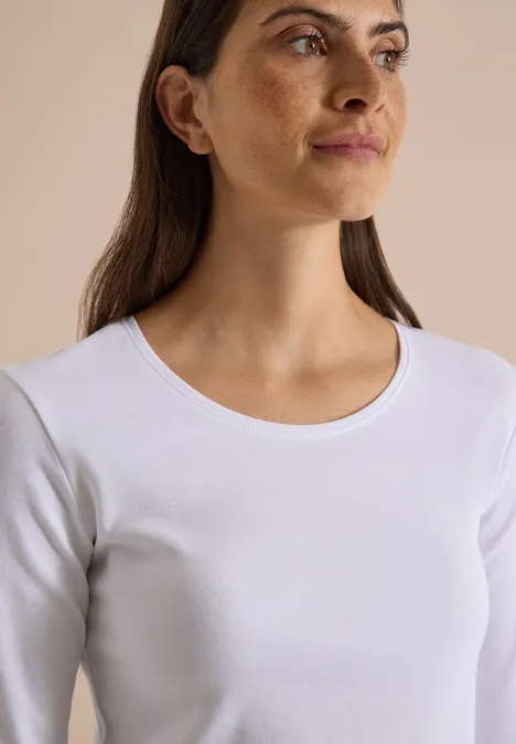 Junge Frau mit braunen Haaren, im weißen Langarmshirt vor neutralem Hintergrund, seitlich blickend.