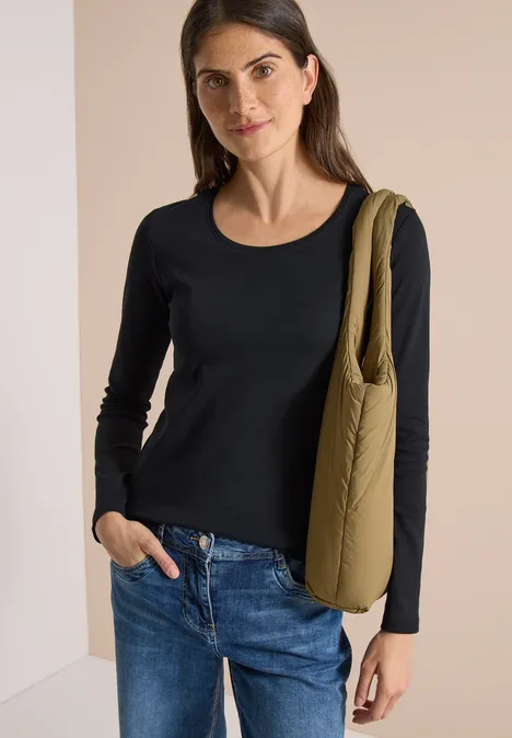 Eine Frau mit schwarzem Langarmshirt, blauen Jeans und olivgrüner Umhängetasche lächelnd