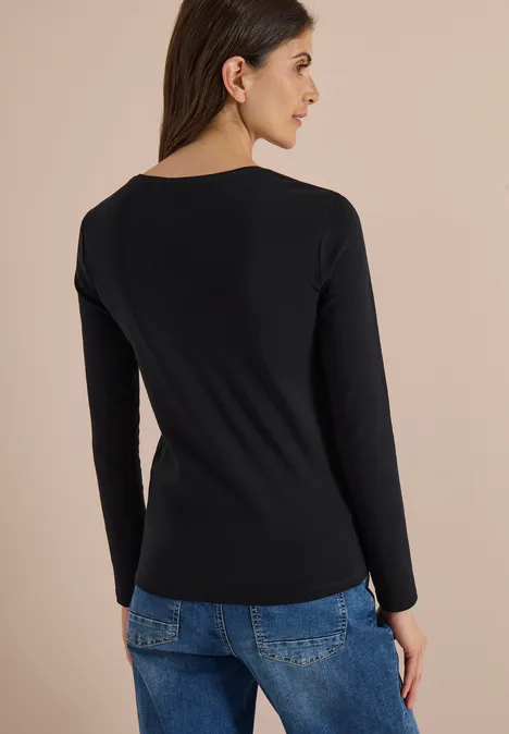 Frau von hinten in schwarzem Langarmshirt und blauen Jeans vor beigem Hintergrund.