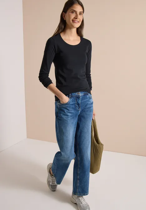 Frau in schwarzem Langarmshirt, blauen Jeans und Sneakern hält eine grüne Tasche und lächelt.