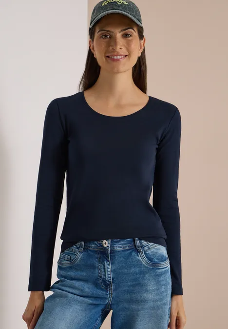 Junge Frau in dunkelblauem Langarmshirt, Jeans und Baseballcap lächelt freundlich.