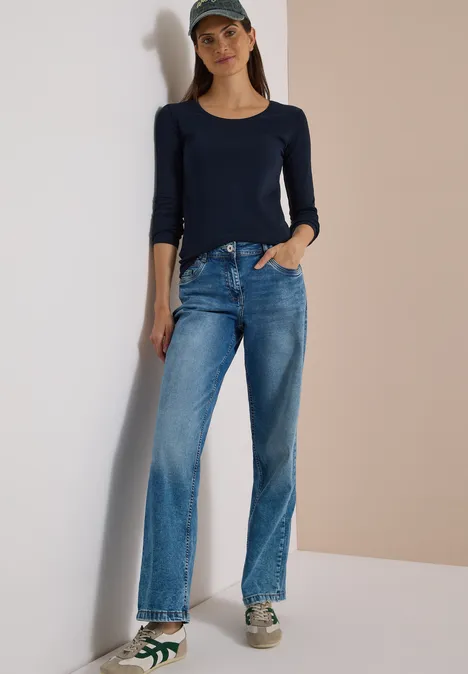 Eine Frau lehnt entspannt an einer Wand und trägt ein dunkelblaues Shirt, Jeans, Baseballcap und Turnschuhe.