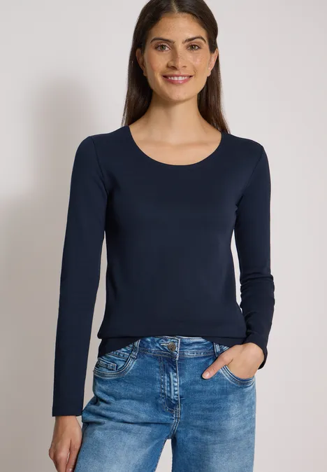 Eine lächelnde Frau trägt eine dunkelblaue Langarmbluse und eine blaue Jeans vor neutralem Hintergrund.