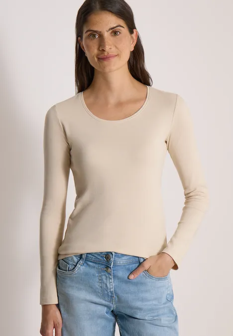 Basic Langarm Shirt mit Rundhals pearl beige Junge Frau in beigefarbenem Langarmshirt und blauen Jeans steht lächelnd mit einer Hand in der Hosentasche.