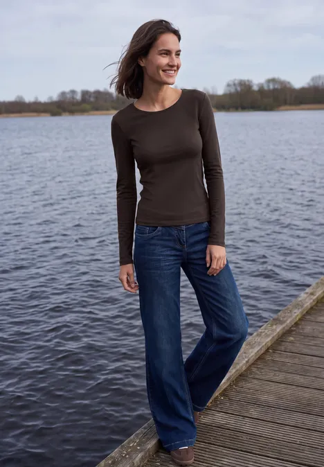 Vrouw in bruin shirt en wijdvallende spijkerbroek die glimlacht op een steiger bij een meer