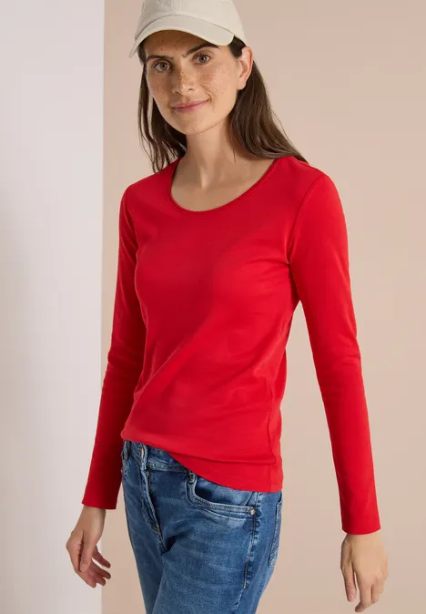 Een vrouw met een rood shirt met lange mouwen, blauwe jeans en een beige pet voor een neutrale achtergrond, met een zachte glimlach.