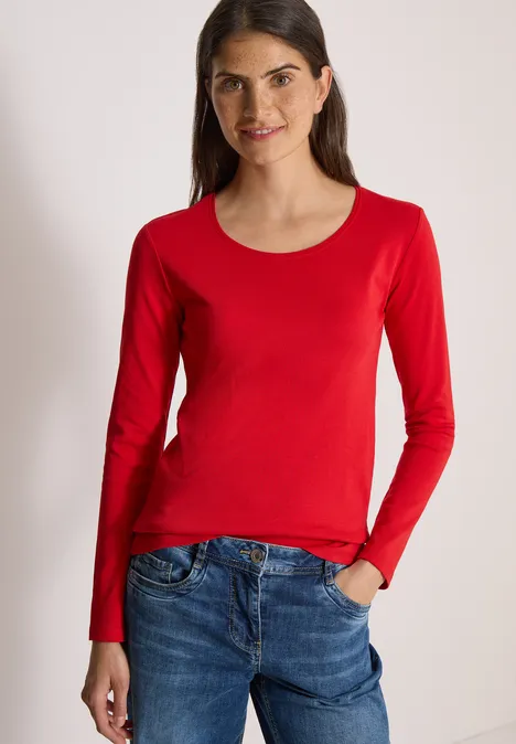 Vrouw draagt rood shirt met lange mouwen en jeans, glimlacht met hand in de zak.