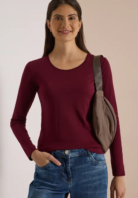 Frau in dunkelrotem Langarmshirt, blauen Jeans und Schultertasche, die lächelnd vor beiger Wand steht