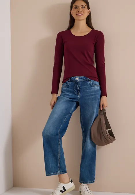 Frau in Bordeaux-Langarmshirt, weitem Jeans, weißen Plateausneakern und brauner Handtasche vor beigem Hintergrund.