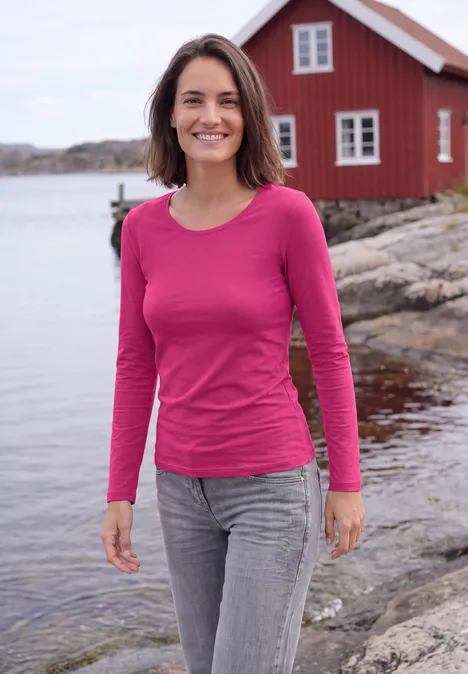 Basic Langarm Shirt mit Rundhals pink flash Frau mit pinkem Oberteil und grauen Jeans lächelt vor rotem Haus am Felsen an der Küste.