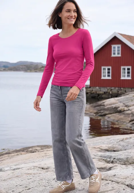 Basic Langarm Shirt mit Rundhals pink flash Eine Frau in rosa Pullover und Jeans spaziert lächelnd am Felsufer vor rotem Haus und Meer.