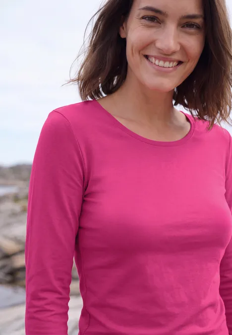 Basic Langarm Shirt mit Rundhals pink flash Fröhliche Frau in langärmeligem pinken Shirt vor unscharfem Hintergrund am Meer.