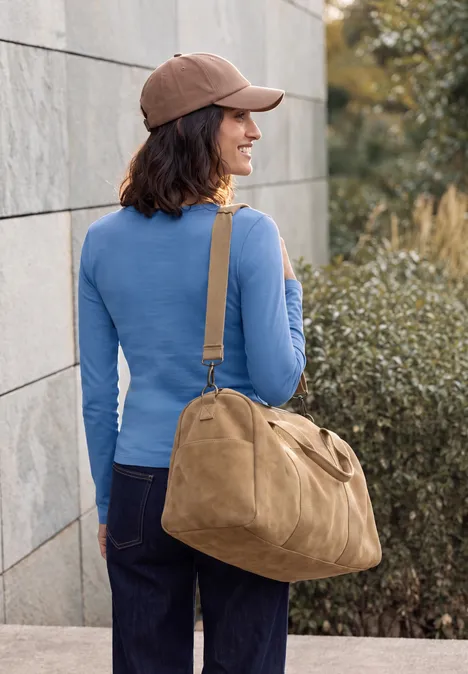 Basic longsleeve met ronde hals north blue Frau in blauem Oberteil, brauner Kappe und Jeans trägt große beige Tasche und steht vor einer Wand.