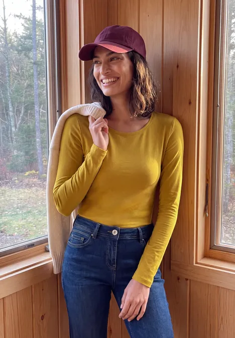 Basic longsleeve met ronde hals golden honey Eine lächelnde Frau im gelben Oberteil, Jeans und roter Mütze hält eine Jacke in einer Holzhütte.