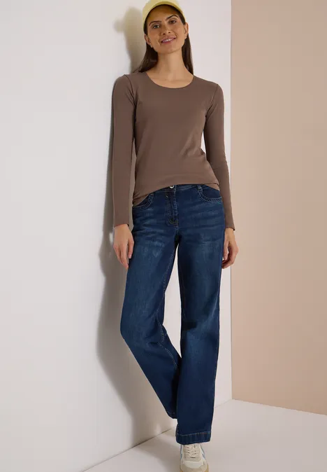 Frau lehnt an Wand, trägt braunes Langarmshirt, blaue Jeans, weiße Schuhe & gelbes Stirnband.