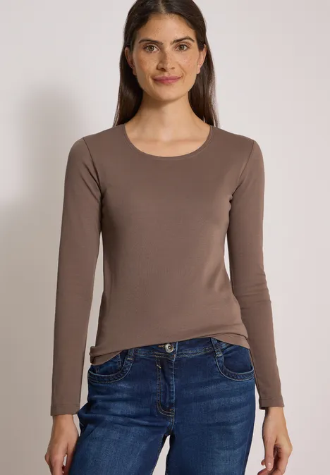 Eine Frau trägt ein braunes Langarmshirt und blaue Jeans und lächelt vor schlichtem Hintergrund.