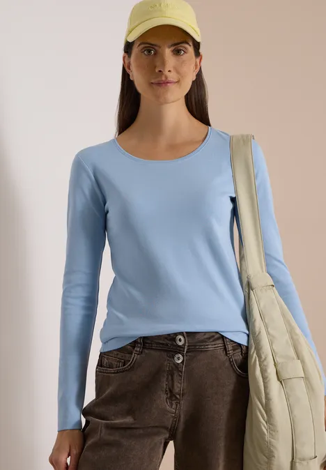 Frau in hellblauem Langarmshirt, brauner Hose, gelber Cap und cremefarbener Stepp-Tasche.