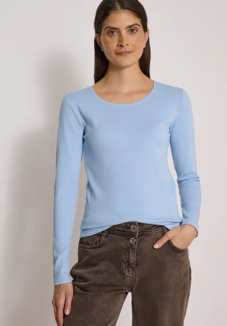 Frau in blauem Langarmshirt und brauner Hose blickt in die Kamera