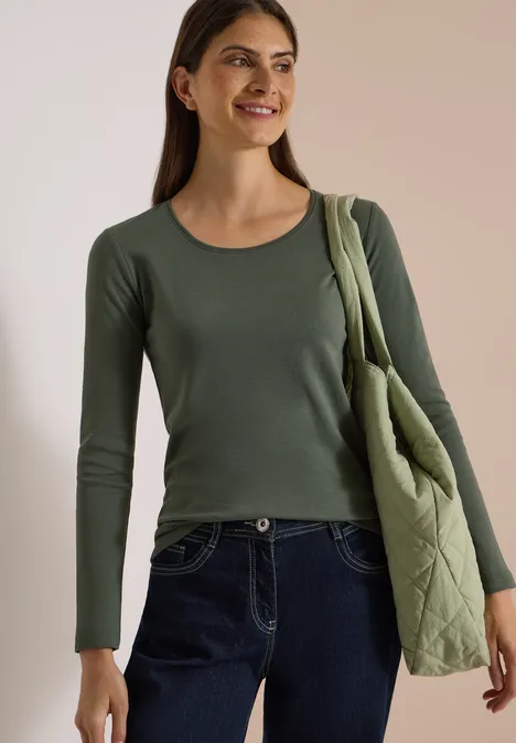 Femme souriante en t-shirt à manches longues vert et sac matelassé assorti devant un fond neutre.