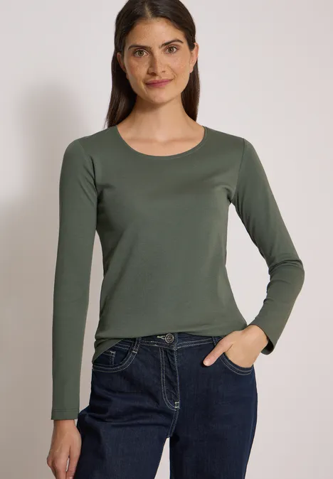 Jeune femme portant un t-shirt à manches longues olive et un jean bleu sur fond neutre.