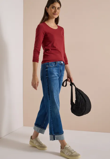 Junge Frau in rotem 3/4-Arm-Shirt, weiten, umgeschlagenen Jeans und beigen Sneakern, mit schwarzer Tasche.