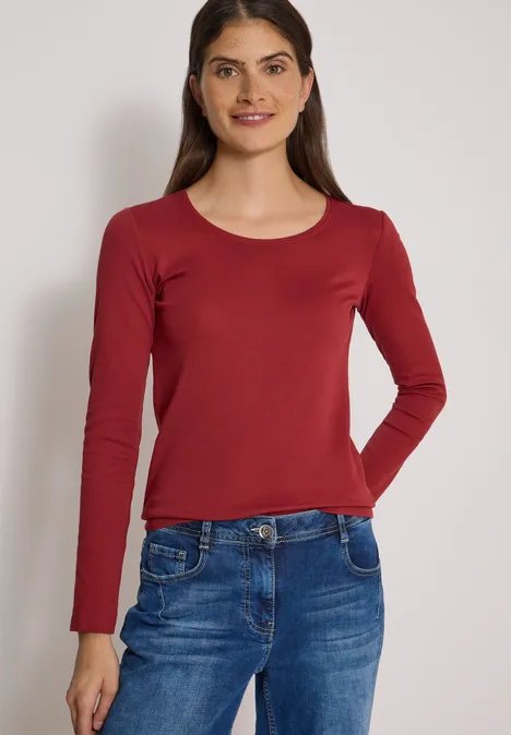 Frau mit langärmligem rotem Shirt und blauen Jeans steht vor hellem Hintergrund.
