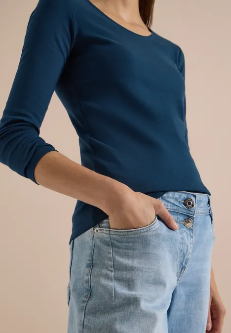 Person trägt ein dunkelblaues Langarmshirt und helle Jeans, eine Hand in der Hosentasche.