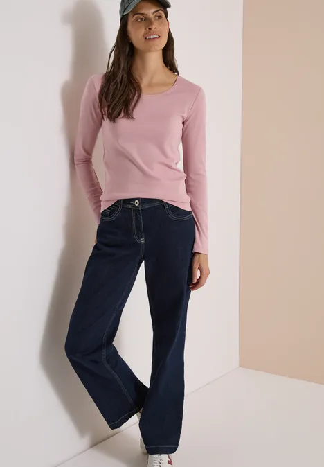 Eine Frau mit rosafarbenem Langarmshirt, weiten Jeans und Cap lehnt an einer Wand.