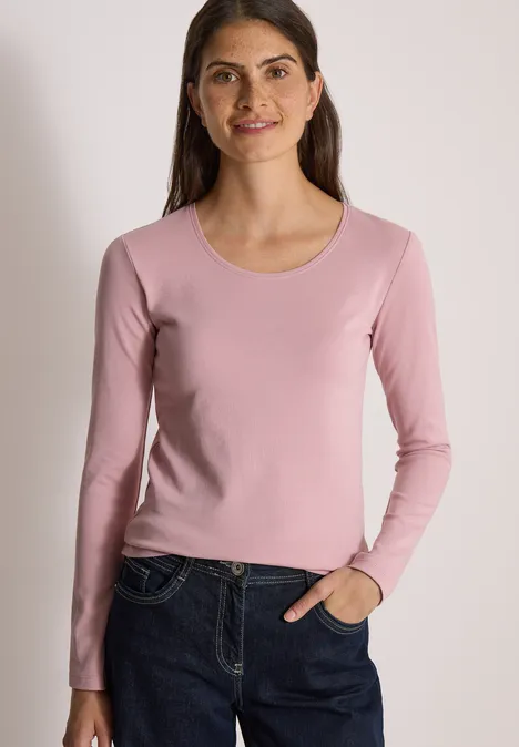 Eine Frau in einem rosa Langarmshirt und Jeans vor hellem Hintergrund.