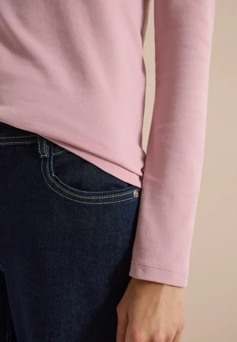 Nahaufnahme einer Person in rosa langem Oberteil und dunkler Jeans, Hand seitlich am Körper.