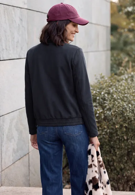 Frau in schwarzer Jacke, Jeans und rosa Cap mit gemusterter Tasche läuft an Mauer und Büschen vorbei.