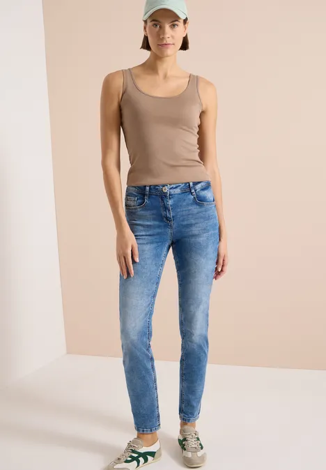 Frau in beige Top, blauen Jeans und gemusterten Sneakers vor neutraler Kulisse.