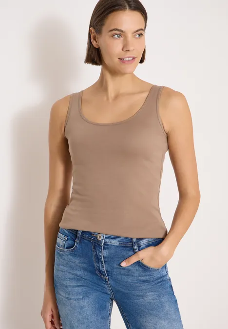 Junge Frau trägt braunes Tanktop und blaue Jeans, eine Hand in der Tasche, vor hellem Hintergrund