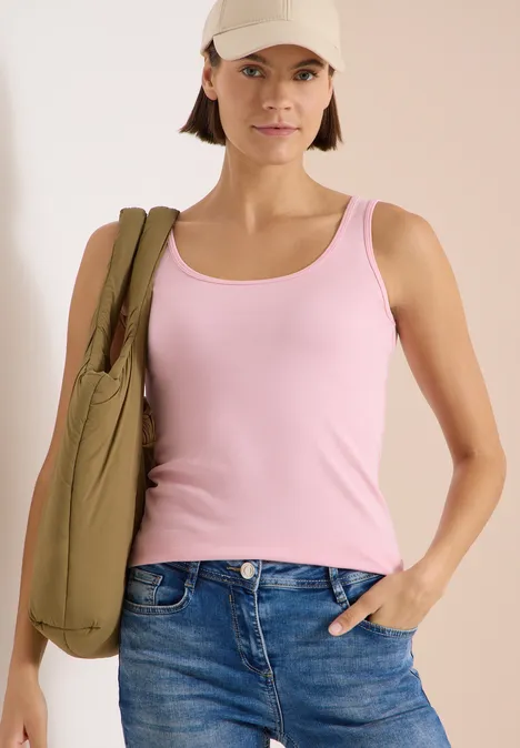 Femme en débardeur rose et jean, sac vert à l’épaule, main dans la poche du jean.