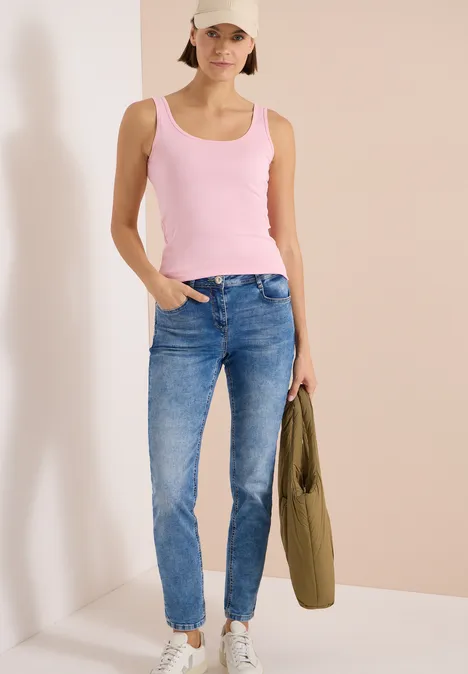 Femme en débardeur rose et jean bleu devant un fond clair, tenant une veste olive.