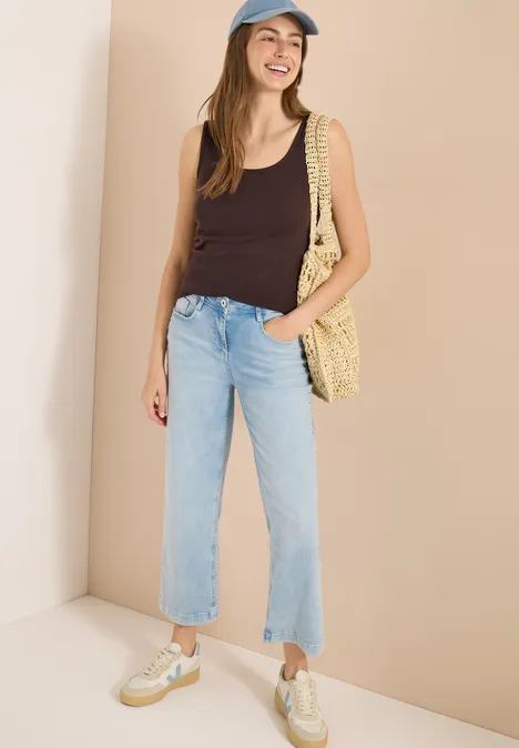 Femme en débardeur noir, jean clair, baskets et sac en osier devant un mur beige.
