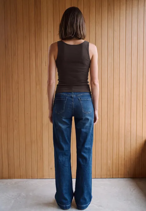Eine Frau steht vor einer Holzvertäfelung, trägt schwarzen ärmellosen Top und blaue Jeans, Rückenansicht.